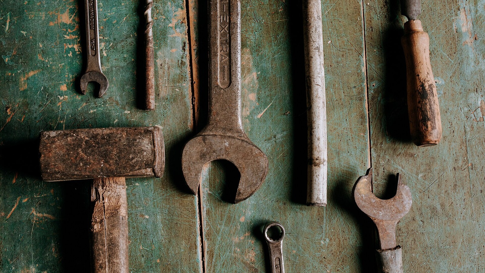 An assortment of hand tools laid out on a green workbench: open-ended and ring spanners, a mallet, a hammer, a drill bit, a file and a chisel.